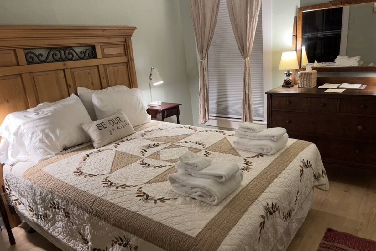 A neatly made bed with a quilt, pillows, and folded towels. There is a wooden headboard, a bedside table with a lamp, and a dresser with a mirror.