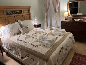 A neatly made bed with a quilt, pillows, and folded towels. There is a wooden headboard, a bedside table with a lamp, and a dresser with a mirror.
