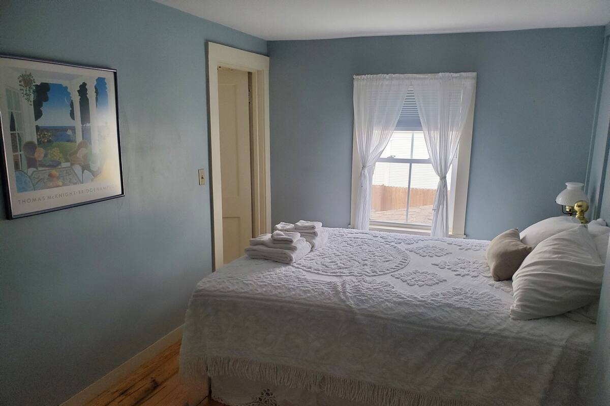 A bedroom with a bed, a window with curtains, a door, and a framed picture on the wall.