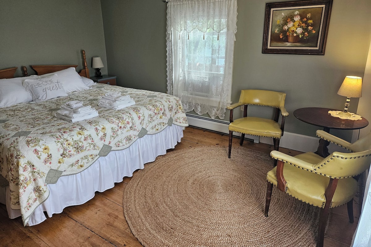 A neatly arranged bedroom with a bed, a chair, a small table, and a painting on the wall.