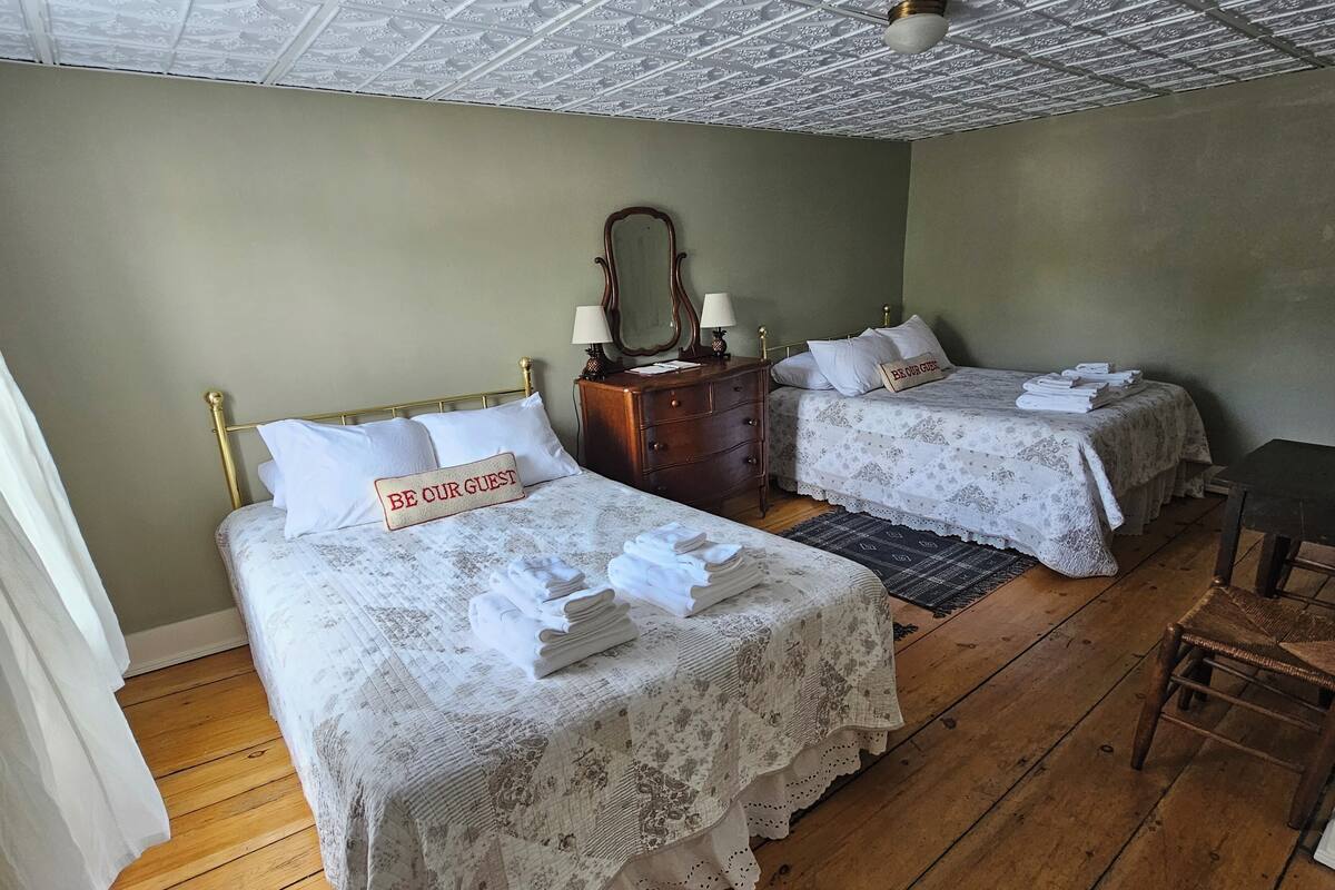 A room with two beds, each with a 'BE OUR GUEST' sign, a wooden dresser, a mirror, and a small table.