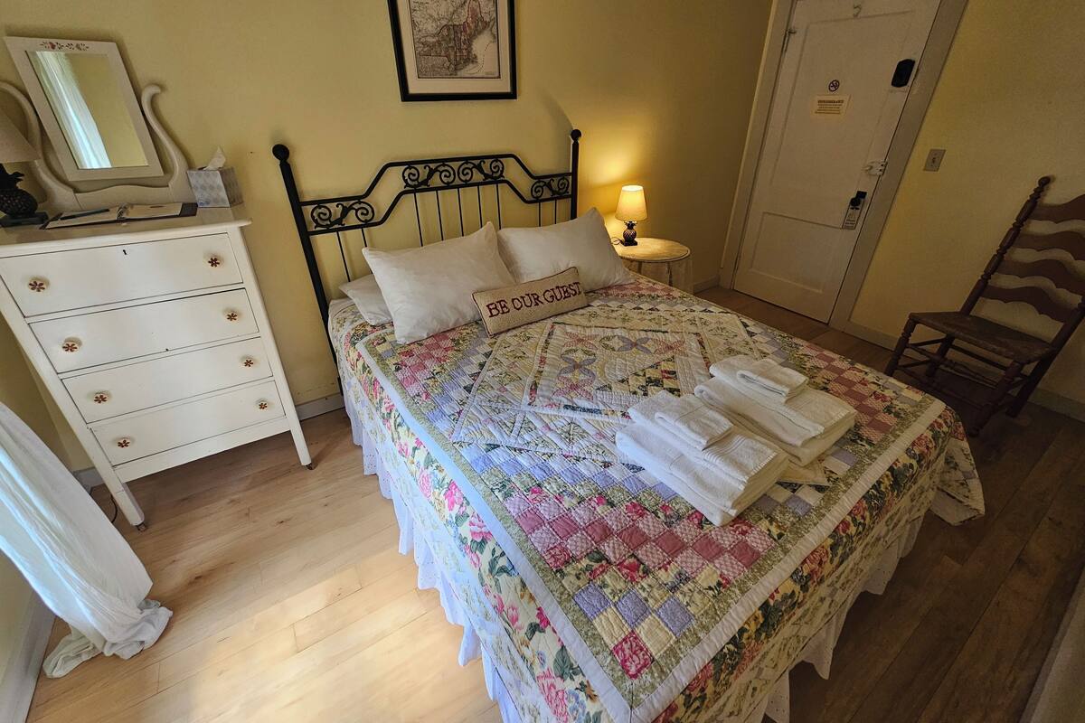 A neatly made bed with a quilt, a bedside lamp, a dresser with drawers, a mirror, and a framed picture on the wall.