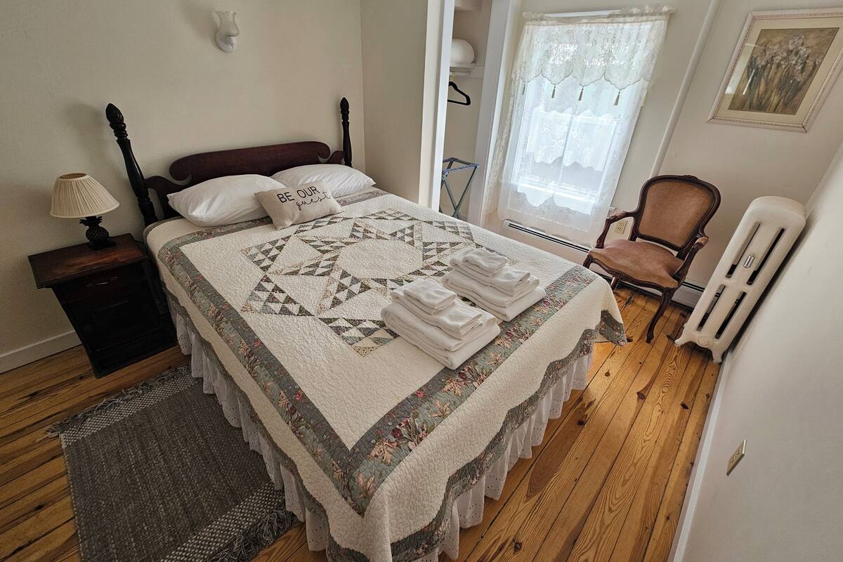 A neatly made bed with a quilt, a bedside lamp, a chair, and a radiator.