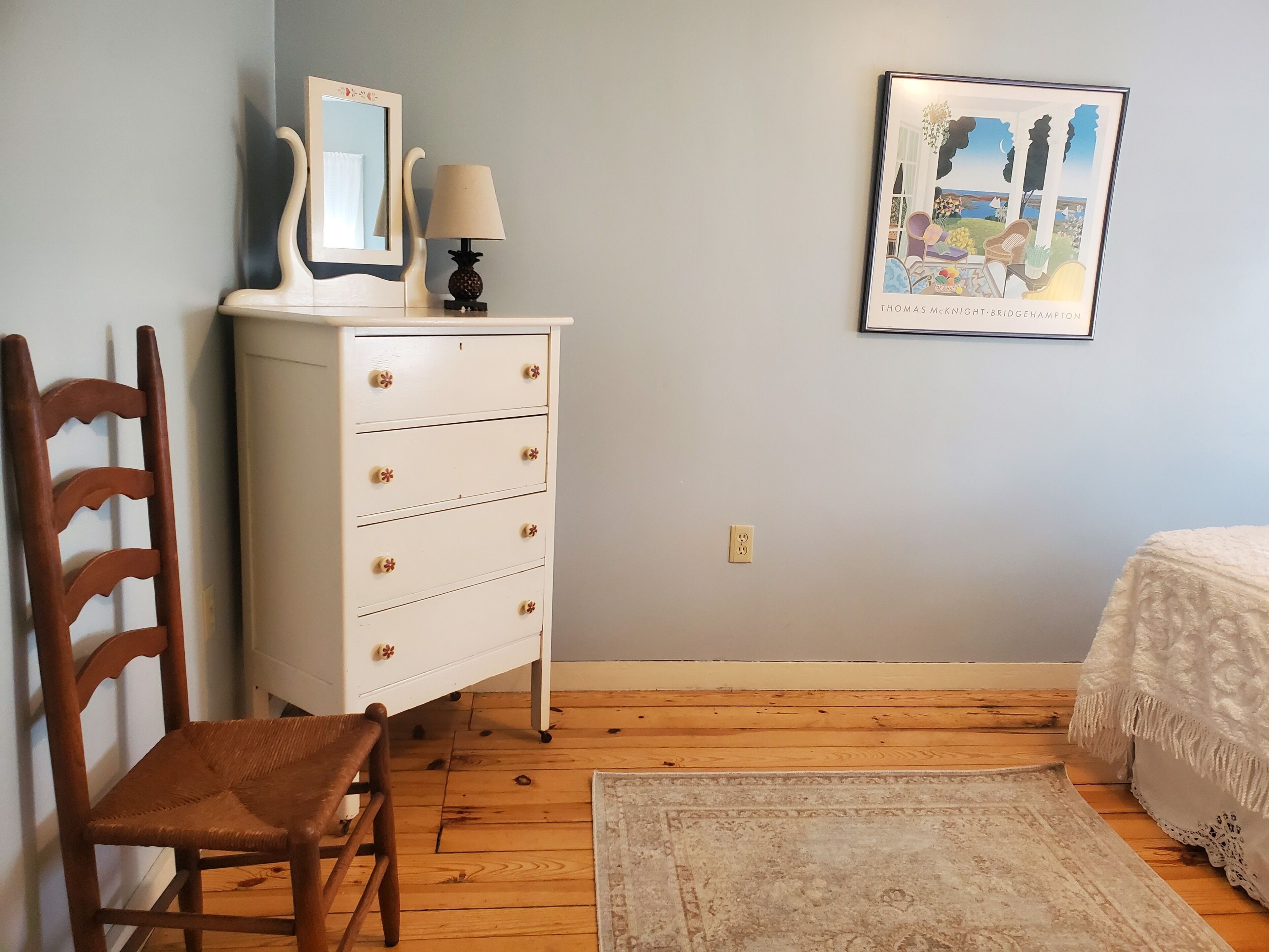 A bedroom with a wooden dresser, a chair, a bed with a white bedspread, a framed picture on the wall, and a wooden floor.