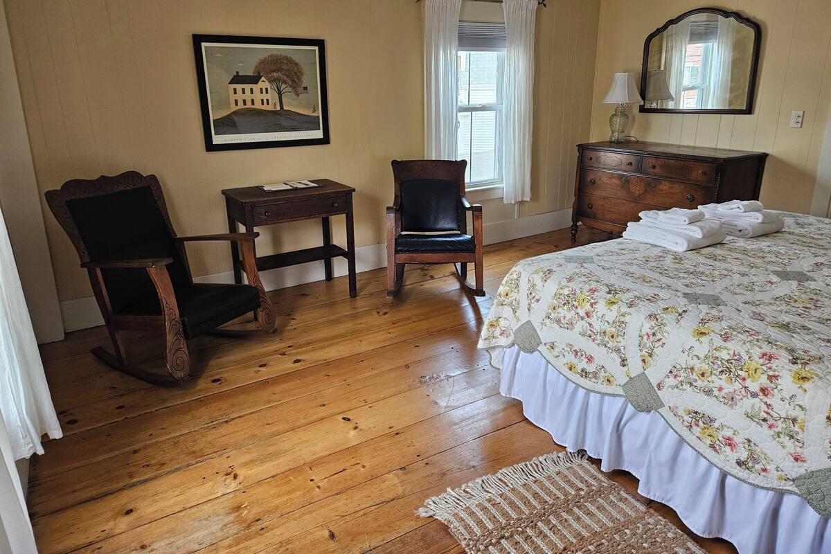 A room with a wooden floor, a bed with a floral bedspread, a rocking chair, a small table, a dresser, a mirror, and a framed picture on the wall.