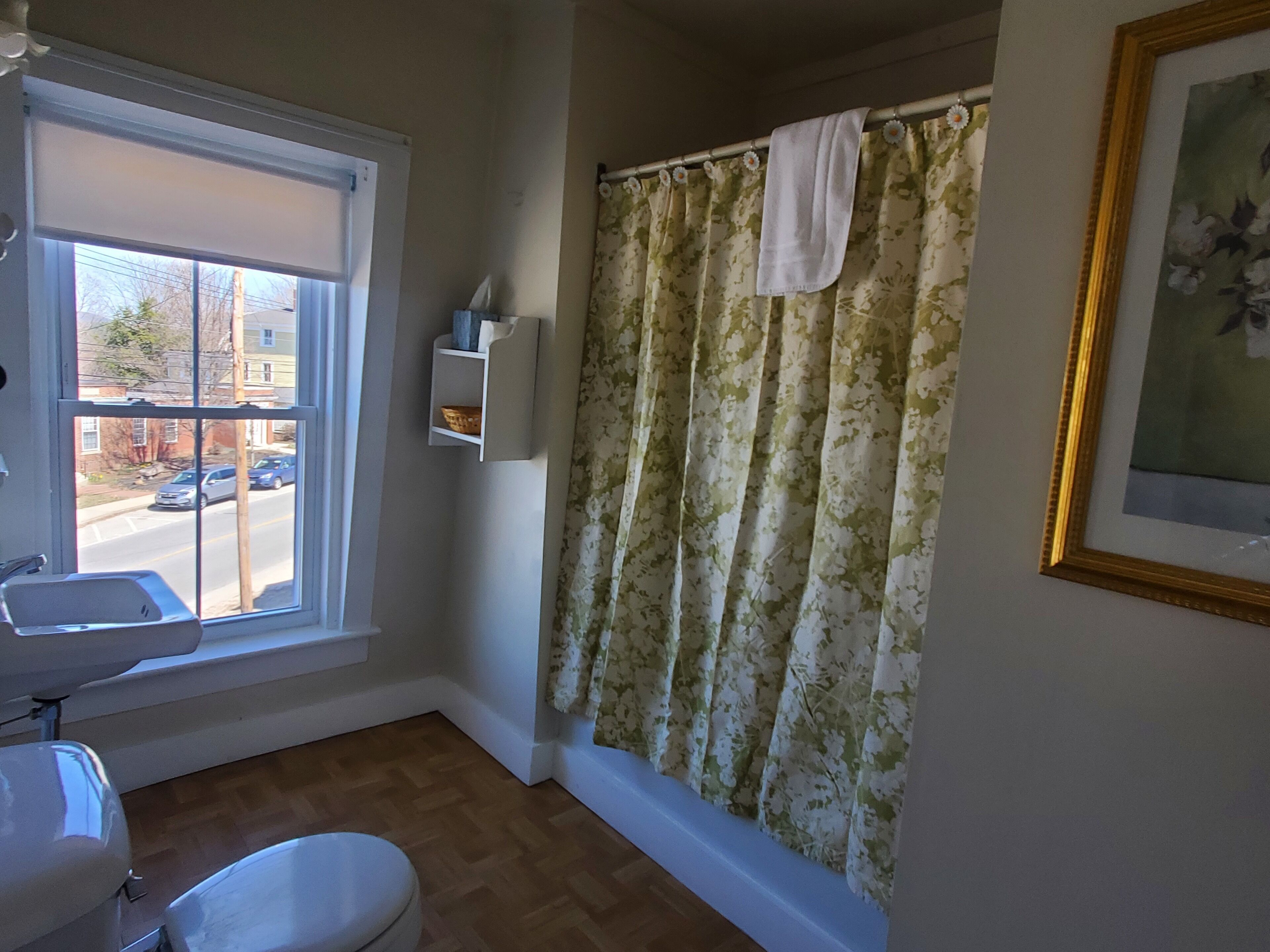 A bathroom with a toilet, sink, window, shower curtain, and a framed picture on the wall.
