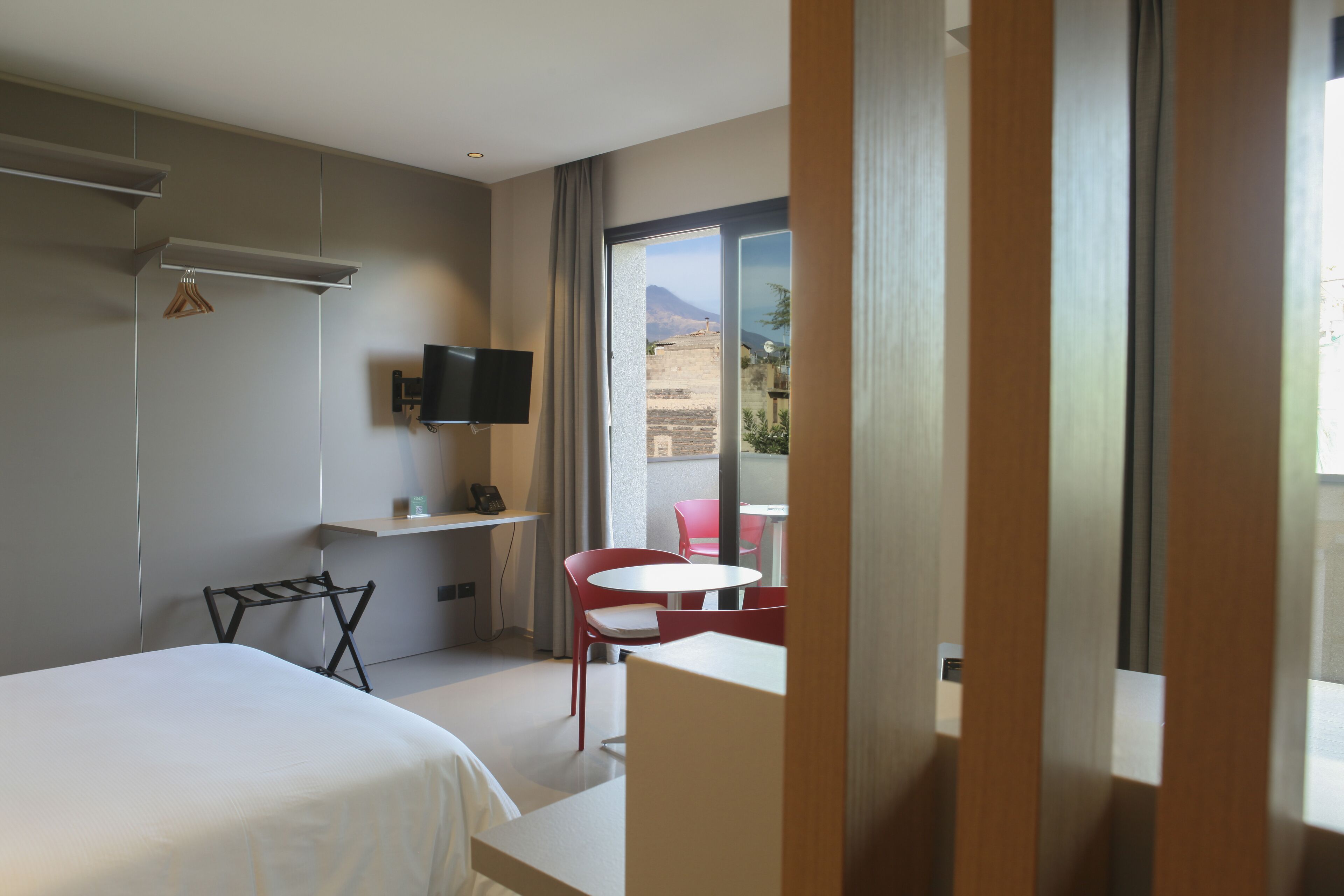 Habitación de hotel con cama, televisor montado en la pared, mesita y silla, y balcón con vistas a las montañas.