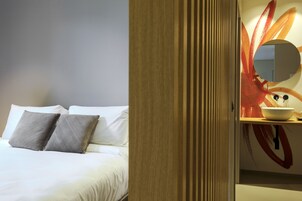 A hotel room with a bed, pillows, a wooden door, and a wall with a circular mirror and abstract art.