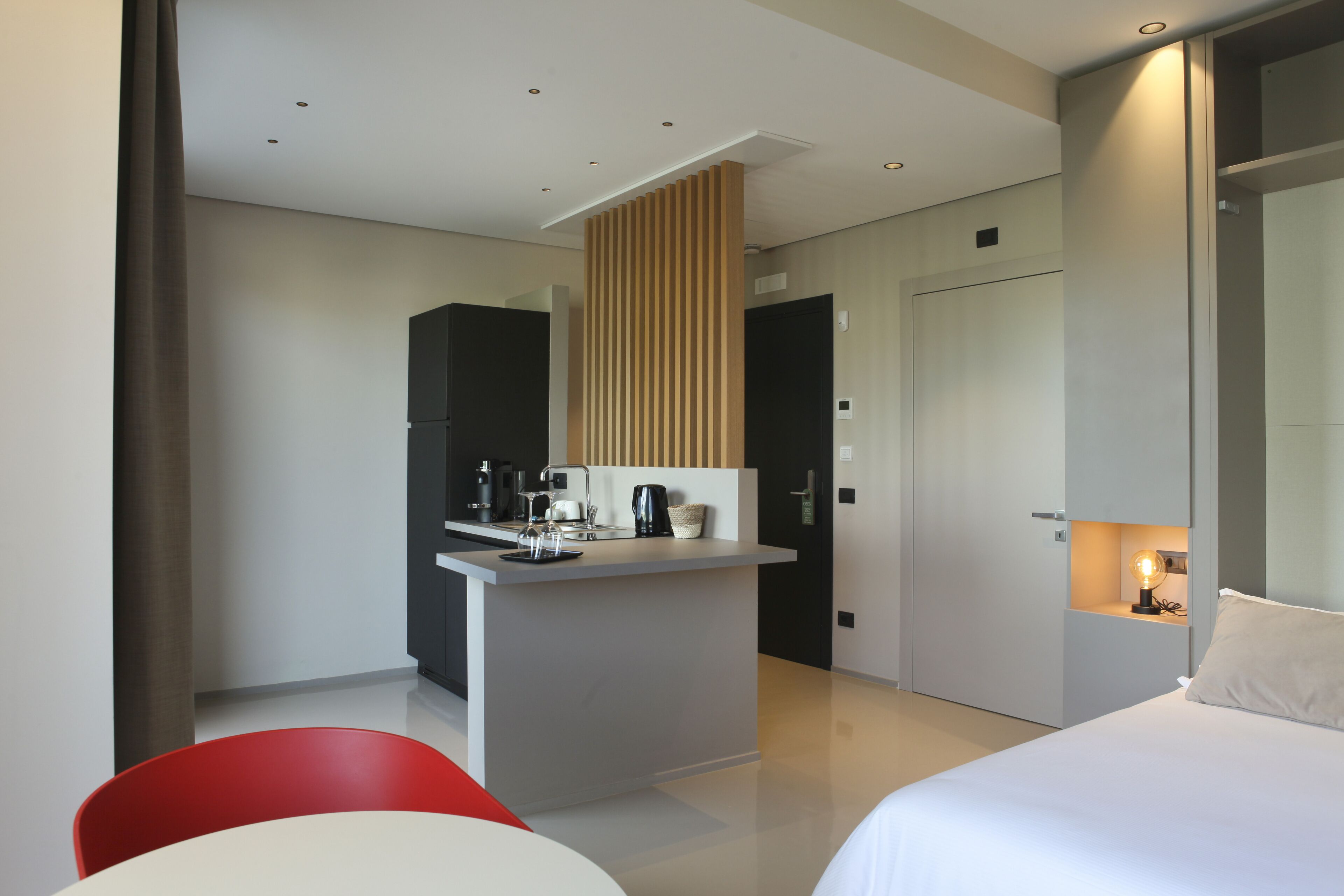 A modern hotel room with a bed, a small kitchenette, a red chair, and a desk.