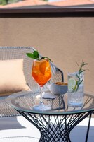 Un bicchiere di bevanda arancione con menta su un tavolino di vetro, un bicchiere d'acqua con limone e rosmarino, e una ciotola di snack.