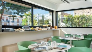 A modern dining area with round tables and green chairs, large windows with a view of buildings and trees, and a buffet with various food items. - Ollen apartments (Catania)