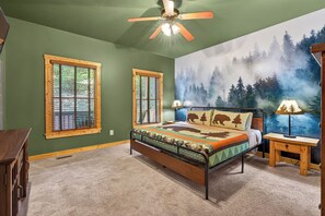 A bedroom with a bed, two windows, a ceiling fan, and a forest-themed wall mural.