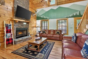 A log cabin interior with a fireplace, leather sofa, and a mountain mural.
