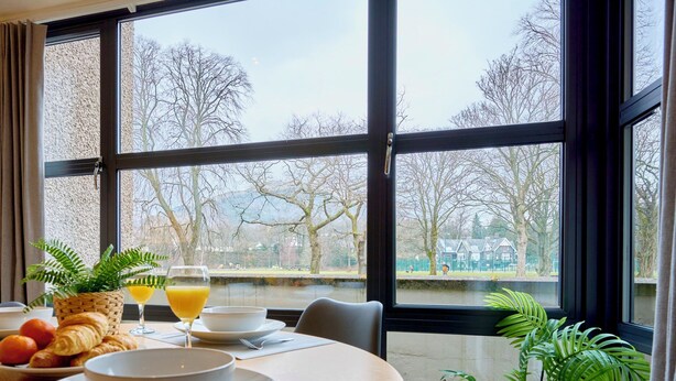 A dining area with a table set for breakfast, a view of trees and a body of water through large windows. - Riverside Park 2 (Keswick)