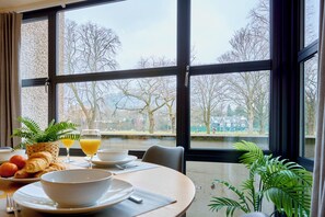 A dining area with a table set for breakfast, a view of trees and a body of water through large windows. - Riverside Park 2 (Keswick)