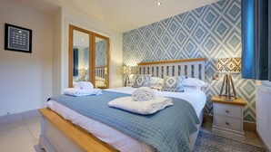 A neatly made bed with a blue quilt, two bedside tables with lamps, a wooden headboard, and a patterned wall. - Riverside Park 2 (Keswick)
