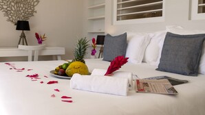 A neatly made bed with a white towel, a fruit plate, and rose petals. There is a magazine on the bed, a lamp, and a decorative wall piece. - Petite Grenade - View, sea access, infinity pool and tennis court (Le Vauclin)