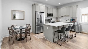 A modern kitchen with grey cabinets, stainless steel appliances, a central island with bar stools, and a dining area with a round table and chairs.
