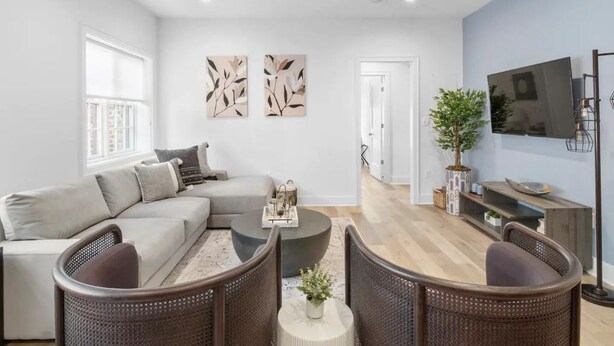 A modern living room with a sectional sofa, a round coffee table, and wicker chairs. There is a flat-screen TV mounted on the wall, a potted plant, and artwork on the wall.