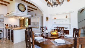 Zona pranzo con tavolo di legno apparecchiato per quattro persone, circondato da sedie. Sopra, un lampadario e sul muro, un orologio.