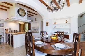 A dining area with a wooden table set for four, surrounded by chairs. A chandelier hangs above, and a clock is mounted on the wall. - Villa for 8 guests with pool and barbecue, YourHouse Son Punta (Felanitx)