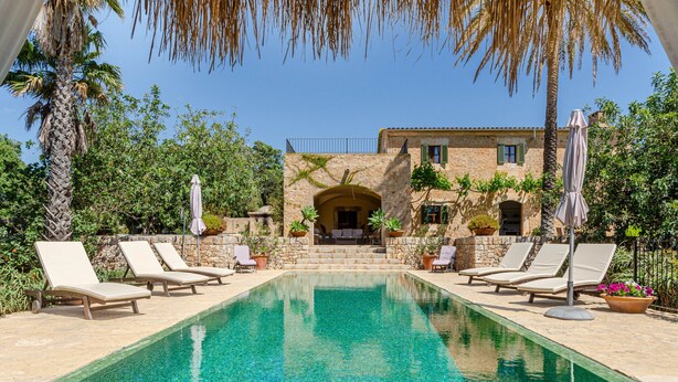 Una piscina con lettini, un edificio in pietra con un balcone e vegetazione circostante.