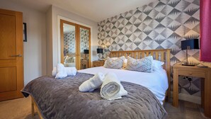 A neatly made bed with a grey duvet, a wooden headboard, and patterned pillows. There are towels on the bed and a nightstand with a lamp. The room has a wooden door and a patterned wallpaper. - Riverside Park 4 (Keswick)