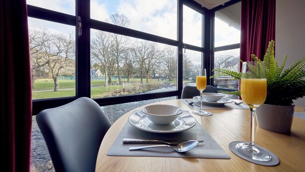 A dining table set with plates, bowls, cutlery, and glasses of orange juice, with a view of a park and trees through large windows. - Riverside Park 4 (Keswick)