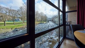A room with large windows overlooking a river, a dining area, and a view of a park with trees and a building in the distance. - Riverside Park 4 (Keswick)