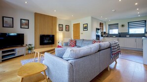 A modern living room with a grey sofa, wooden panel wall, flat-screen TV, and a small wooden table. - Riverside Park 4 (Keswick)