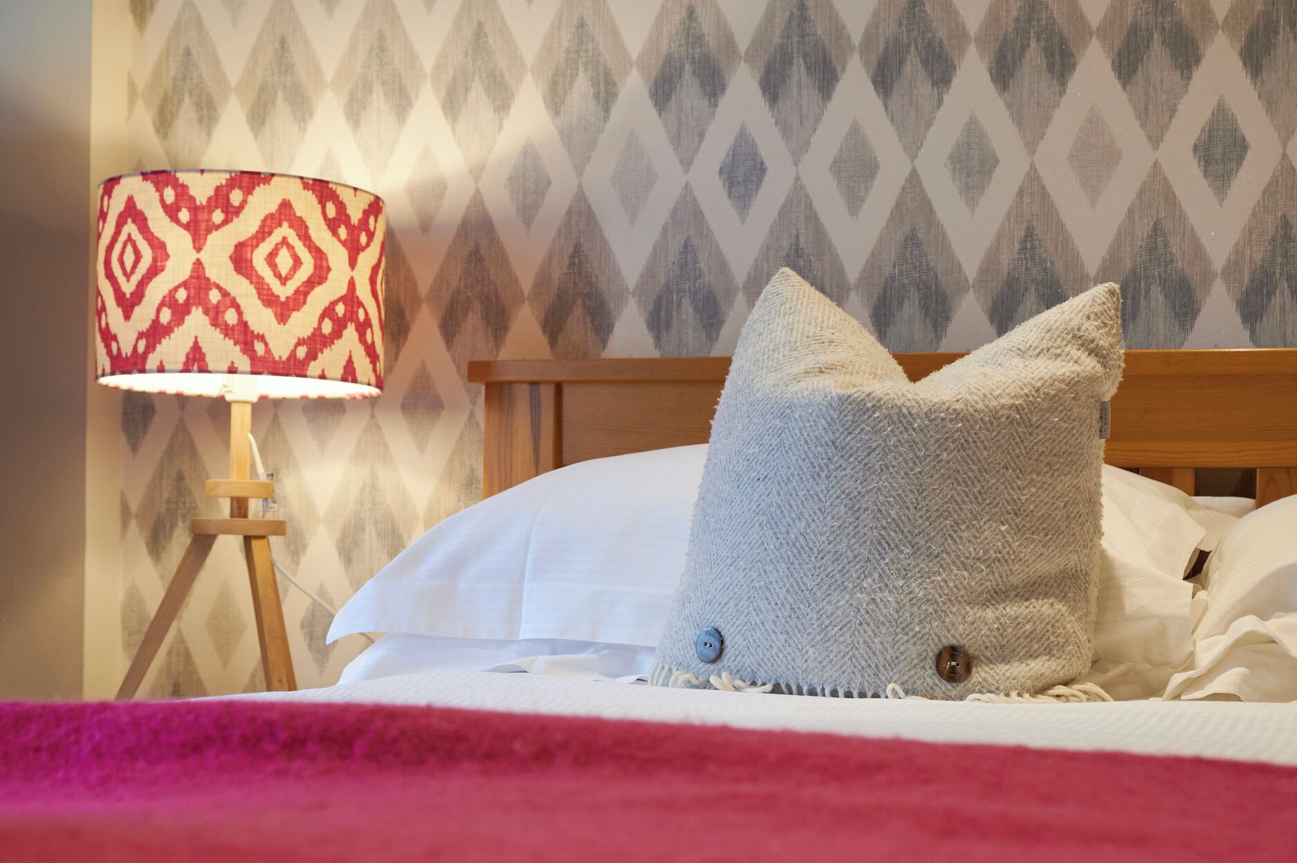 Una cama cuidadosamente arreglada con un almohadón blanco y un almohadón gris texturizado, una lámpara estampada en rojo y blanco sobre un tocador de madera, y un papel tapiz con diseño al fondo.