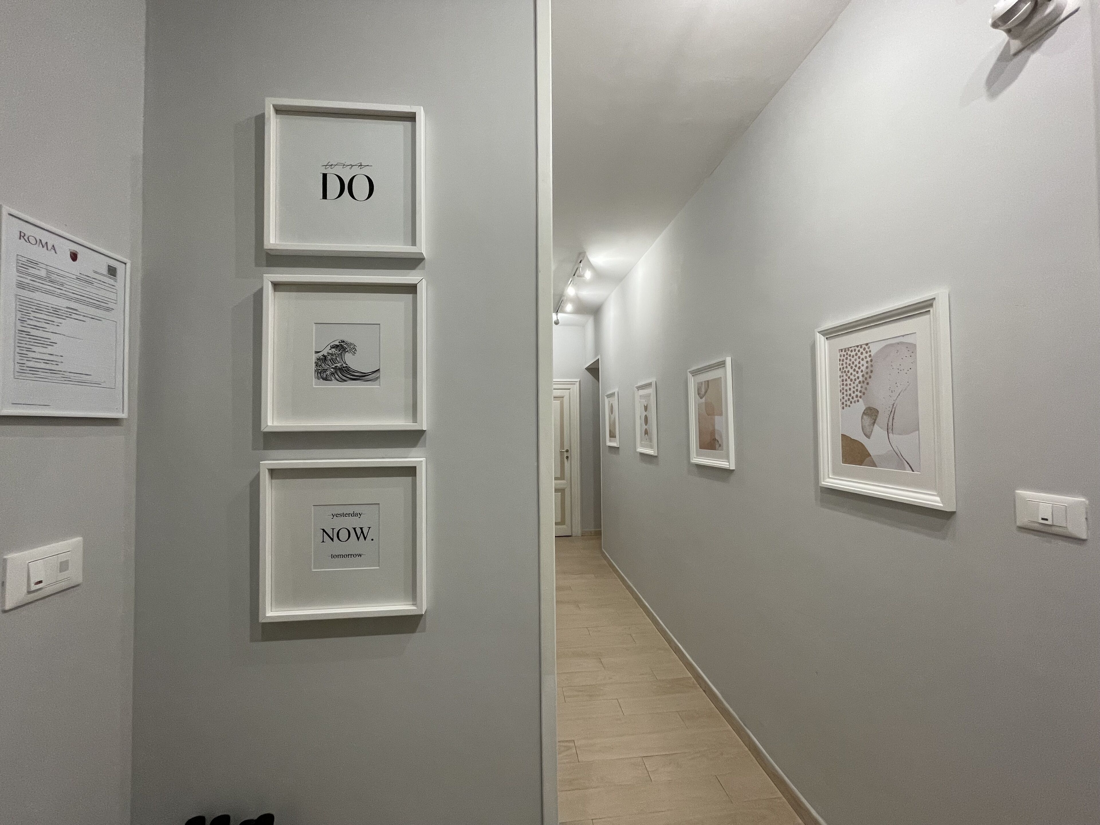 A hallway with framed artworks on the walls, a light switch, and a door at the end.