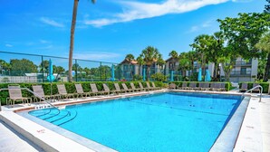 A rectangular swimming pool with clear blue water, surrounded by lounge chairs and umbrellas, and a fence with palm trees in the background. - Lovely 2 Bedroom/ 2 Bath Across Siesta Key Beach! (Siesta Key)