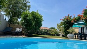 Una piscina azul cristalina con sillas de descanso y sombrillas, rodeada de vegetación y un edificio al fondo.