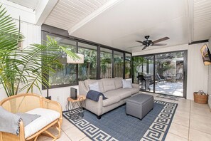 Relax in this spacious living room with a sofa, wicker chair, and ottoman, perfect for unwinding. Enjoy the view from the large windows and step outside to the patio area with barbecue grill.