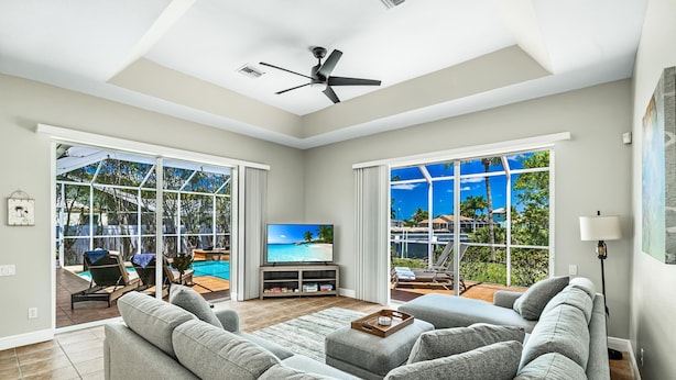 A spacious living room with a large gray sectional sofa, a flat-screen TV, and a ceiling fan. Large windows offer a view of a pool and outdoor furniture. - Pet friendly villa; gulf access canal w/elec heated pool & pretty views! Villa Sienna - SWFL Rentals (Cape Coral)