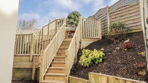 A wooden staircase leading to a deck area with a view of the sky and a garden with plants and mulch.