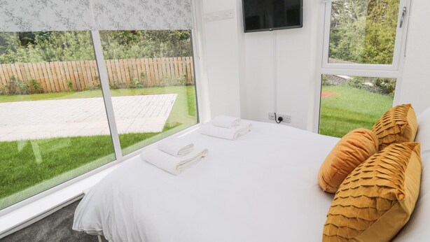 A bedroom with a large bed, two pillows, a TV on the wall, and a view of a garden through the windows.