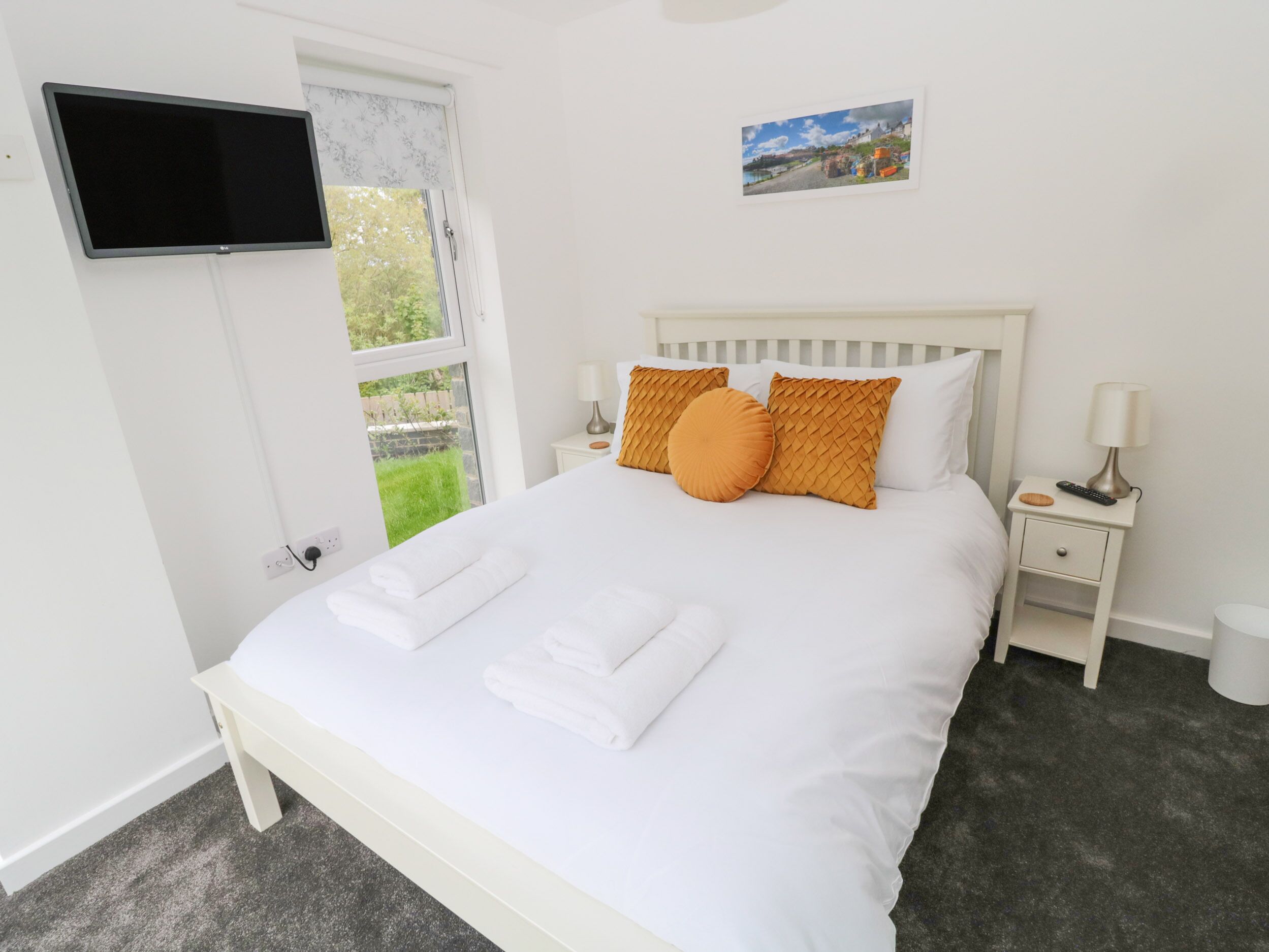 A neatly made bed with white linens and two orange pillows, a flat-screen TV mounted on the wall, a small bedside table with a lamp, and a picture of a scenic landscape above the bed.