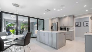 A modern kitchen with grey cabinets, a stainless steel refrigerator, and a dining area with a white table and black chairs. - Stylish & Bright 3-Bedroom Home with Backyard, Bikes & Pool by Esthetic Stays (Naples)