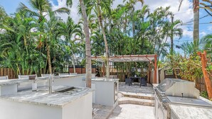 An outdoor kitchen area with a grill, sink, and seating area surrounded by palm trees. - Stylish & Bright 3-Bedroom Home with Backyard, Bikes & Pool by Esthetic Stays (Naples)