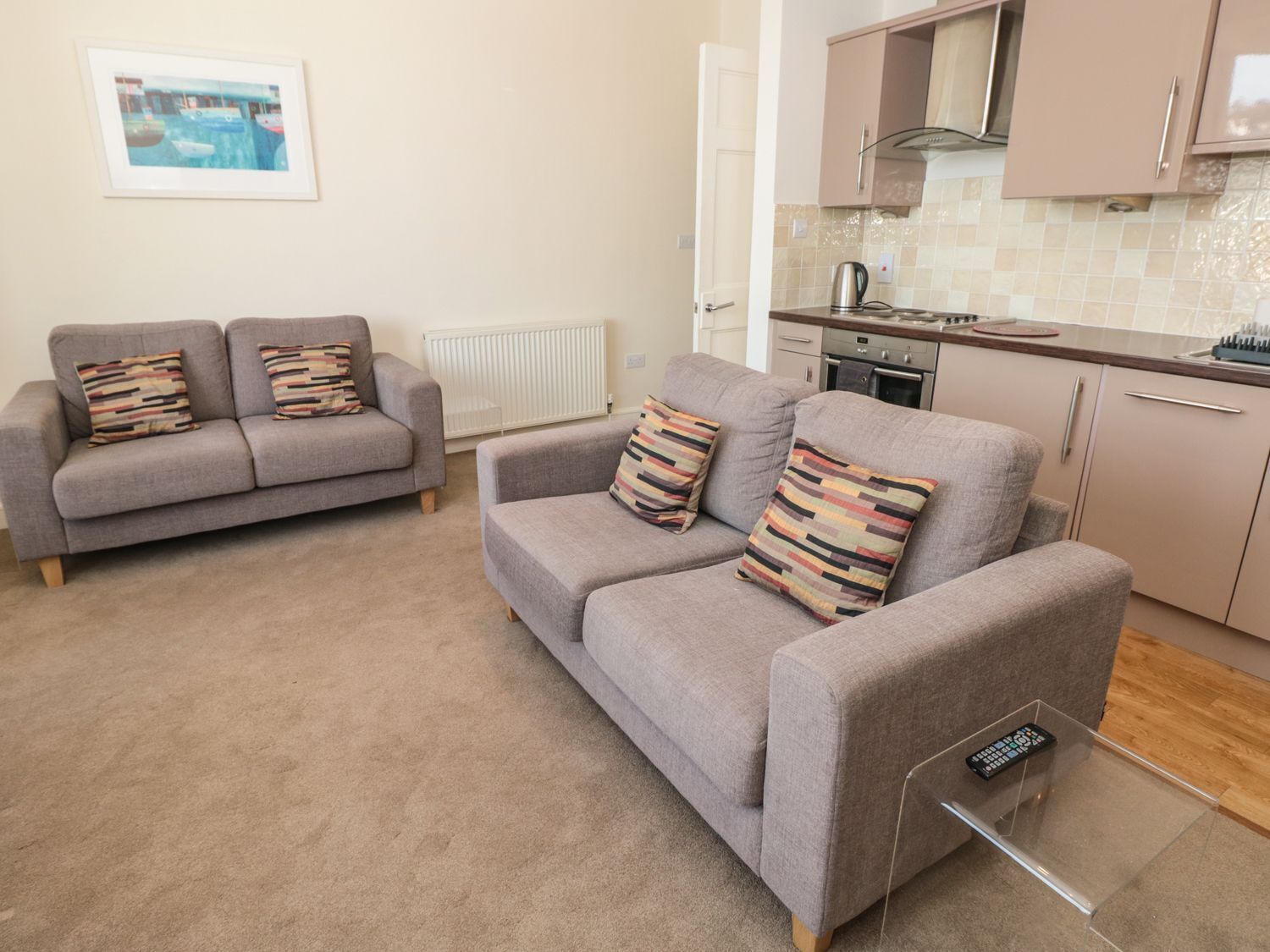 A living room with two grey sofas, each with striped cushions, a small coffee table with a remote control, and a kitchen area with cabinets and a stove.