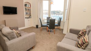 Una sala de estar con dos sofás, un televisor, una estantería, una mesa de comedor con sillas y vista al mar.