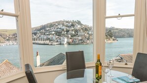 Un comedor con mesa de vidrio, sillas, una botella de vino y una copa. Un libro está sobre la mesa. La habitación tiene ventanas amplias con vistas a un puerto y edificios al otro lado del agua.