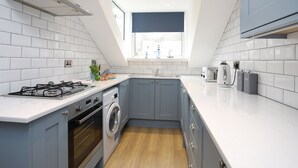 A modern kitchen with white tiles, a gas stove, a washing machine, and a window with a view. - Anchors Rest (Y Felinheli)
