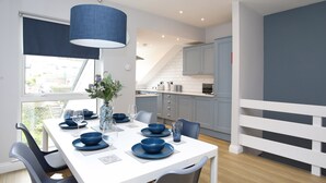 A modern kitchen with white cabinets, a dining area with a white table set for six, and a view of the outside. - Anchors Rest (Y Felinheli)