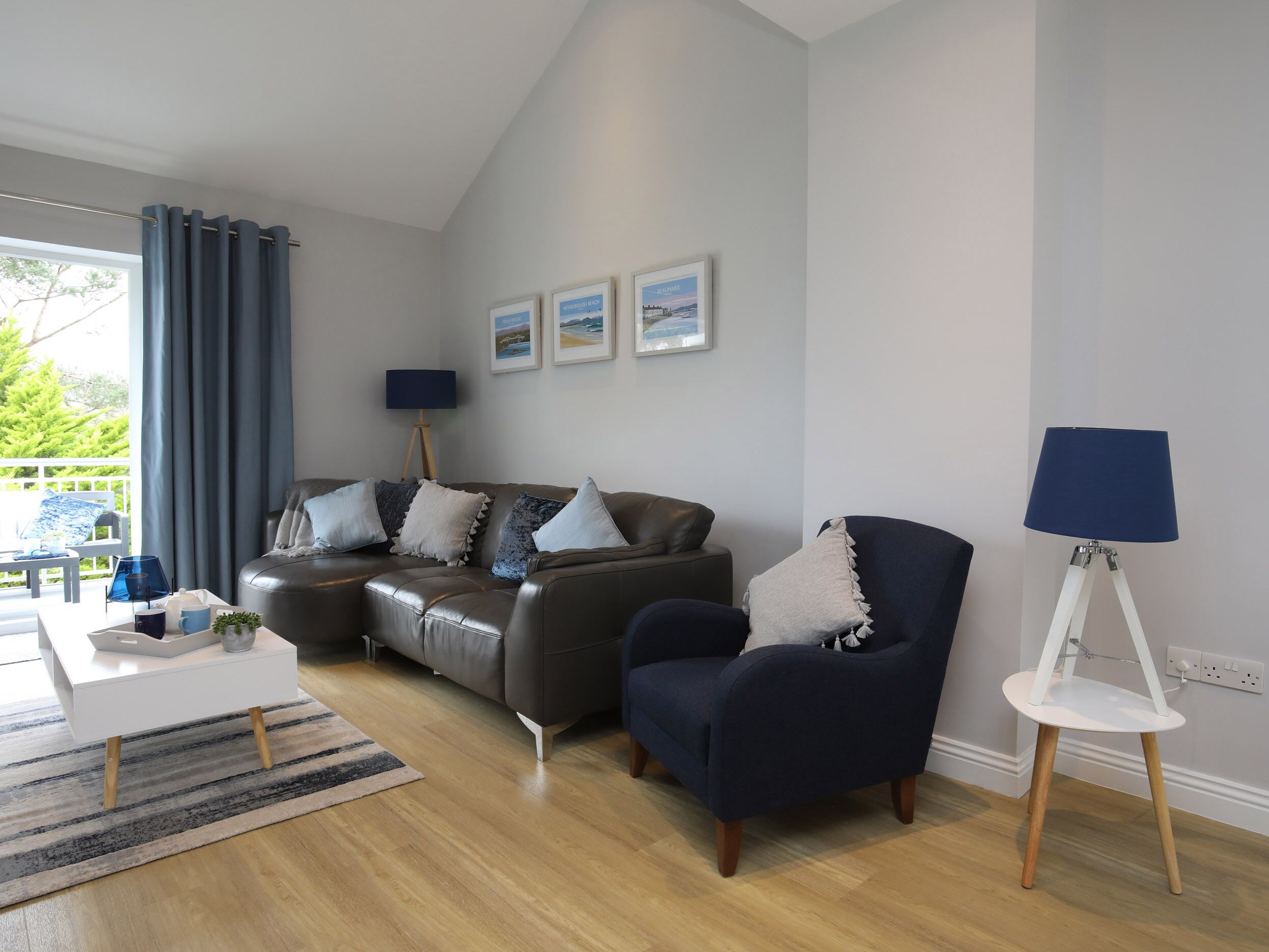 Un salon moderne avec un canapé gris, un fauteuil foncé et une table d’appoint blanche. Trois tableaux encadrés ornent le mur, ainsi qu’une lampe sur pied et une fenêtre avec des rideaux.