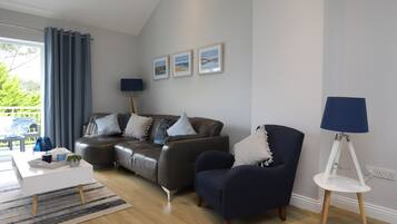 Un salon moderne avec un canapé gris, un fauteuil foncé et une table d’appoint blanche. Trois tableaux encadrés ornent le mur, ainsi qu’une lampe sur pied et une fenêtre avec des rideaux.