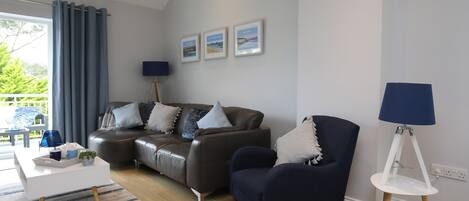 A modern living room with a grey sofa, a dark armchair, and a white side table. There are three framed pictures on the wall, a floor lamp, and a window with curtains.