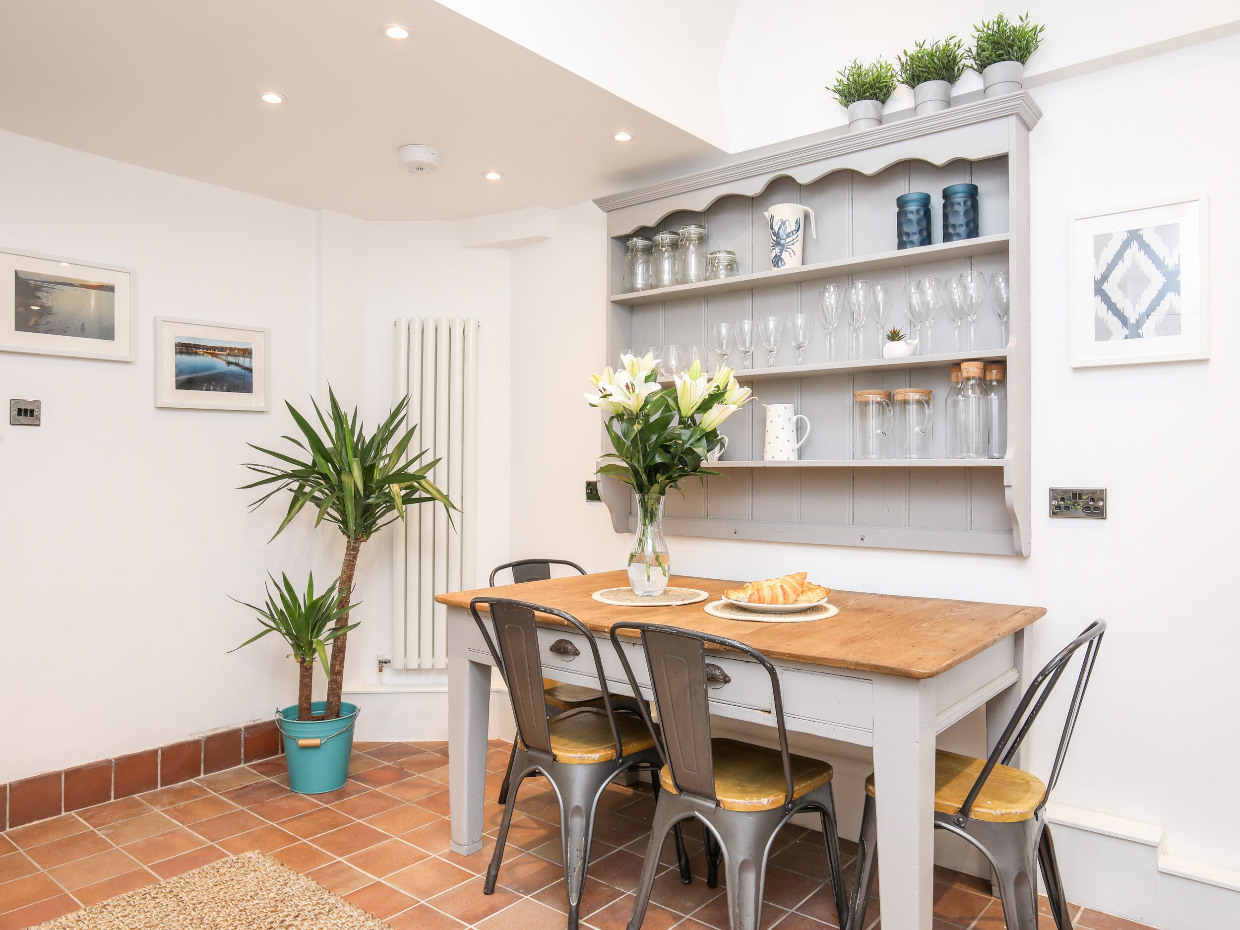 Un comedor con una mesa de madera, sillas metálicas, un florero con flores y una estantería con copas y frascos.
